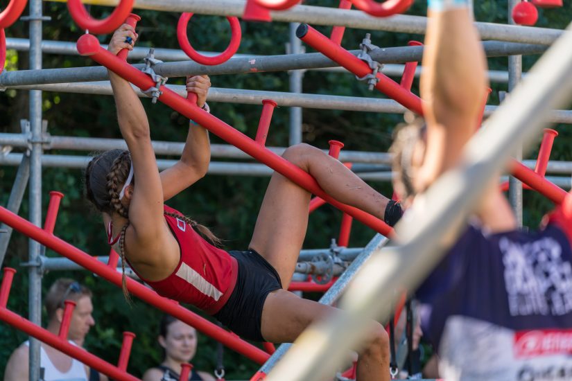 A woman, dressed in athletic gear, climbs a jungle gym as part of a competitive obstacle course. Other participants are visible in the foreground and background.