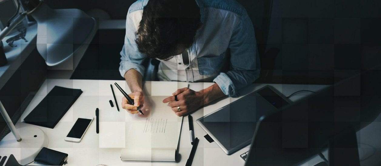 Man drawing on a sheet of paper at a desk in front of a computer