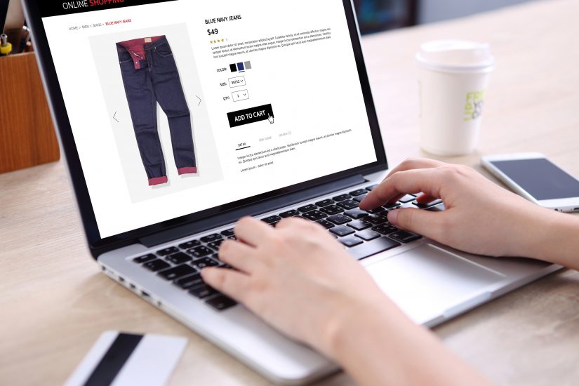 Consumer buying blue navy jeans on an eCommerce website with a smartphone, credit card, and coffee on a wooden desk.
