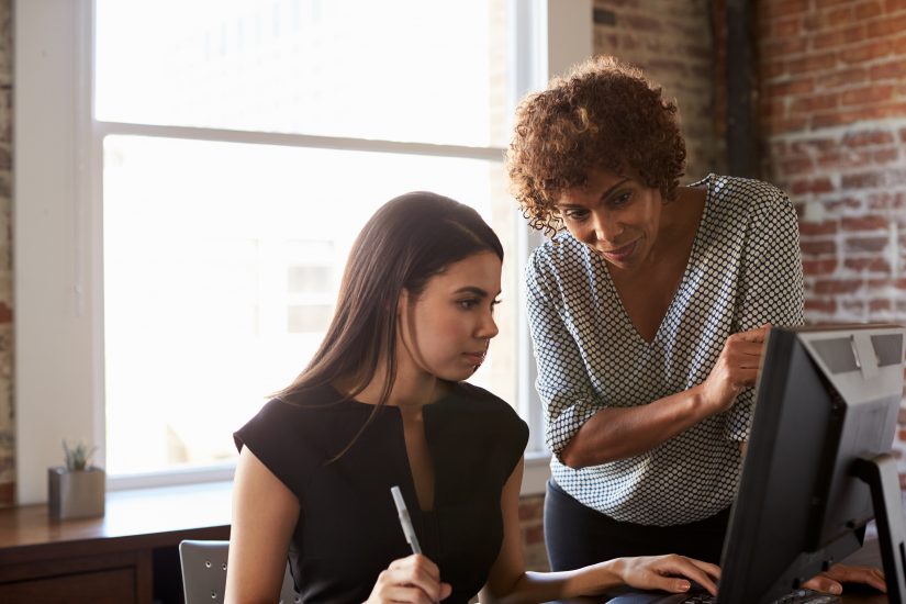 A woman looks over the should of another woman, who is sitting in front of a computer. The woman at the computer is holding a pen while moving the mouse with the other hand.