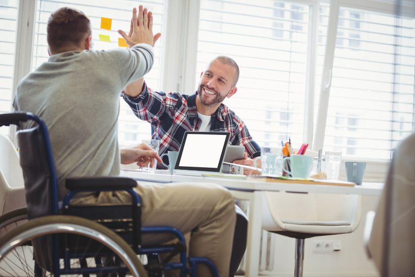 Man in wheelchair high-fives individual over a computer. 