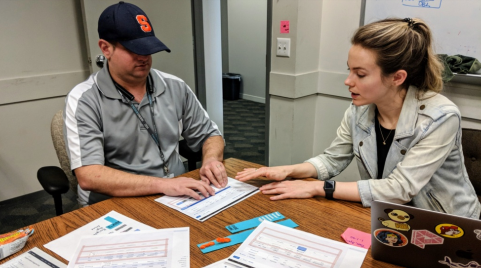 A woman works with a visually impaired man who uses braille to read a design system. 