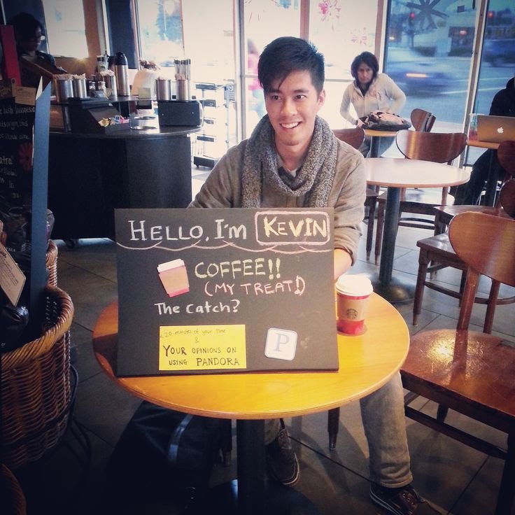 A researcher sits behind a sign recruiting coffee shop customers for a hallway usability test.