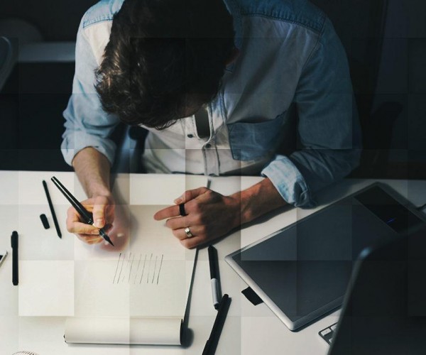 Man drawing on a sheet of paper at a desk in front of a computer